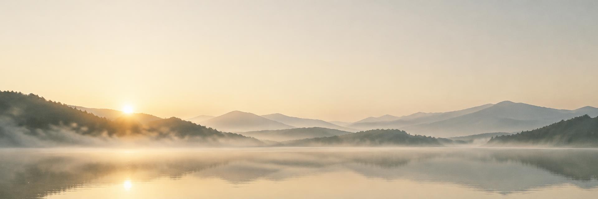 Soft mist drifting over a calm mountain lake as the sun rises behind forested hills